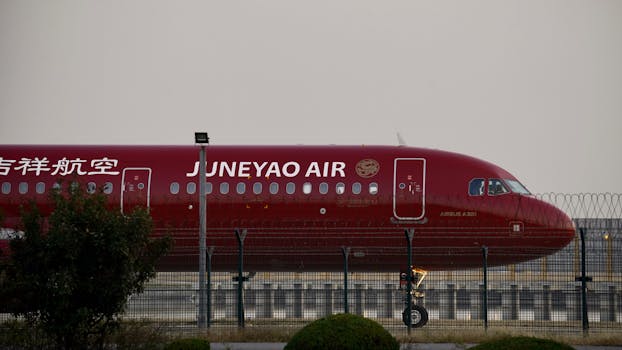 Juneyao Air Airbus A321 in red livery parked behind a fence at an airport.