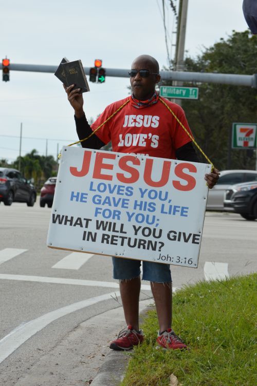 Free A man holds a religious sign at a busy intersection, promoting faith with a Bible in hand. Stock Photo