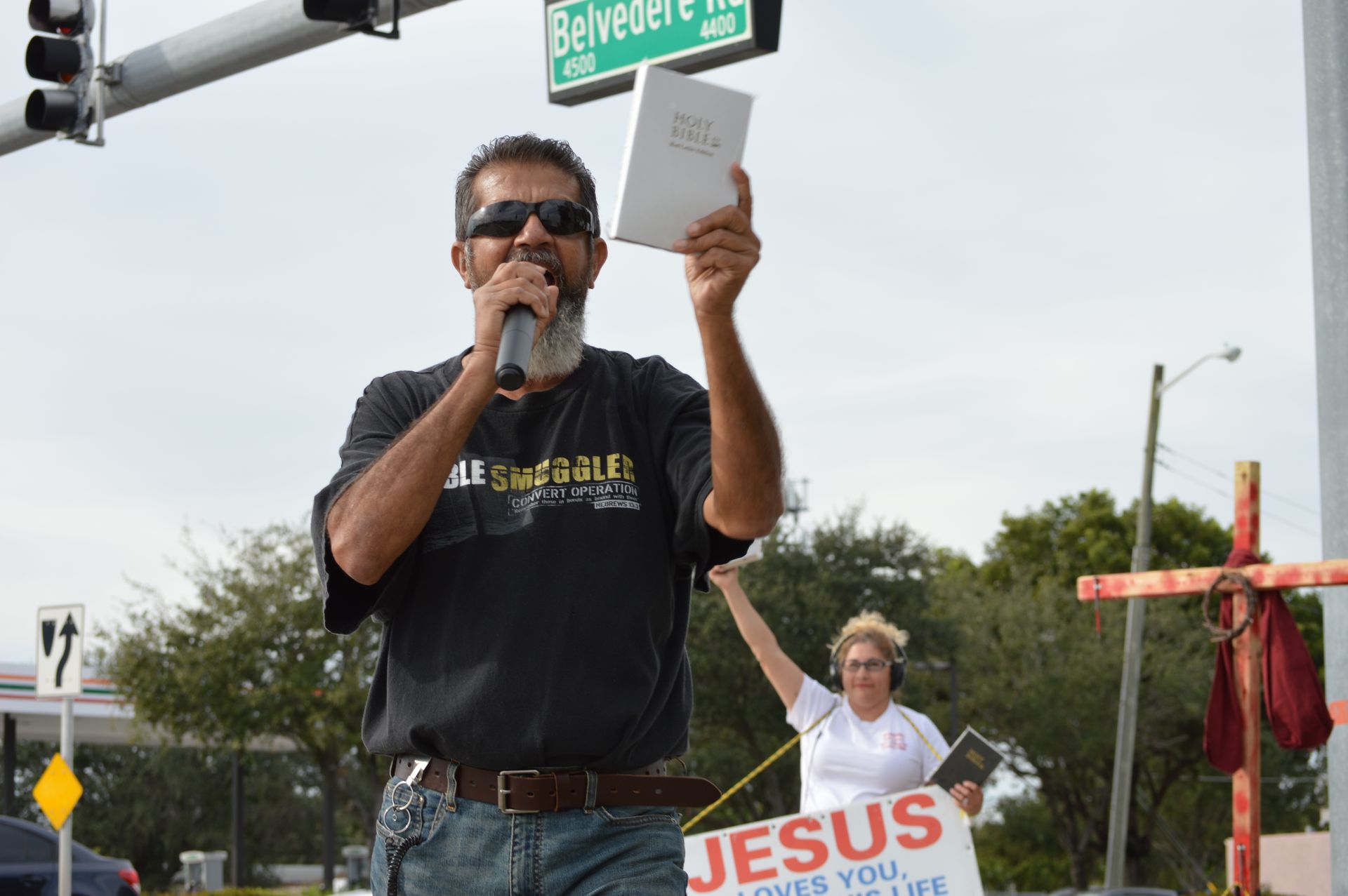 Man holding a Bible and speaking into a microphone during an outdoor religious gathering.