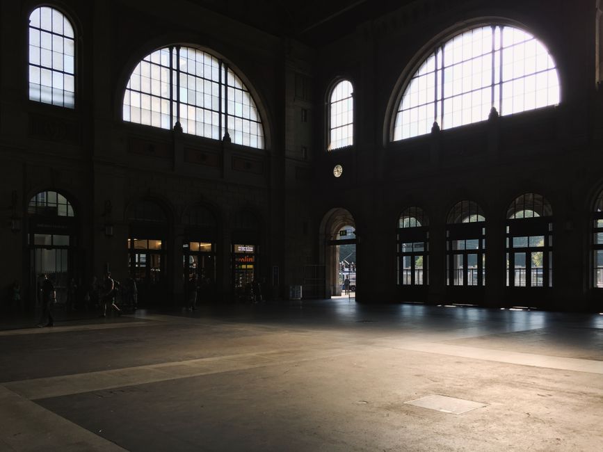 Sunlit interior of a spacious building hall with large arched windows casting shadows.