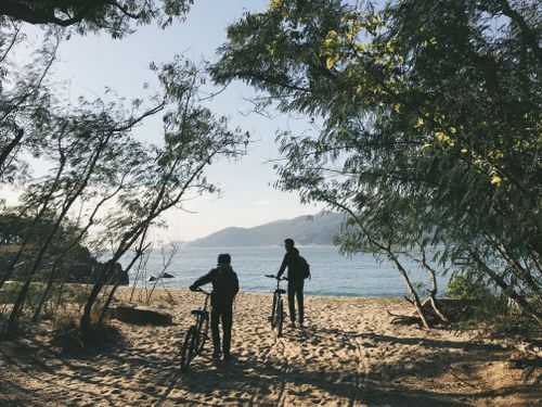 Free Two cyclists walking with their bikes on a sandy beach path surrounded by trees and scenic coastal views. Stock Photo