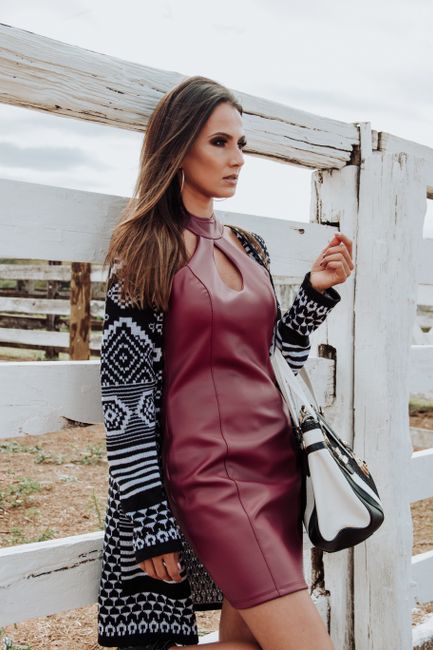 Stylish woman in a burgundy dress and patterned coat standing by a wooden fence, showcasing modern fashion.