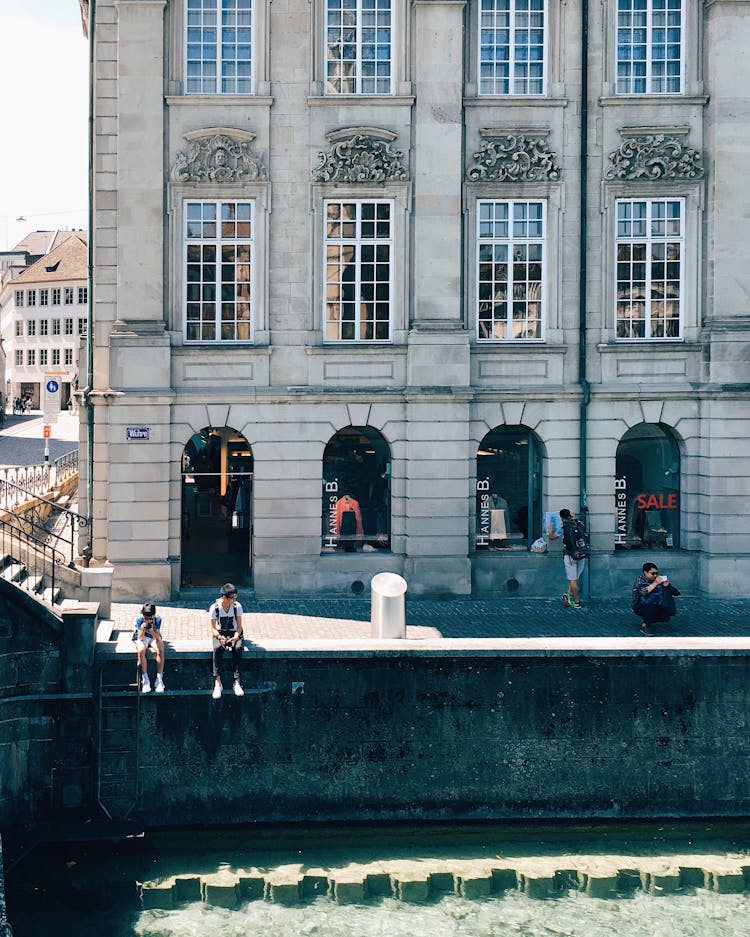 Boys Sitting On A Wall By A River In A City 