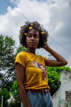 A young woman with flowers in her hair stands confidently outdoors under a cloudy sky.