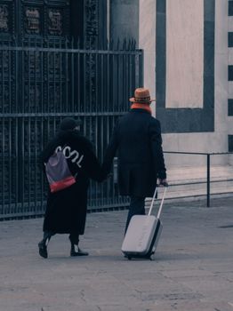 A stylish couple walking together, holding hands with luggage, in an urban environment.