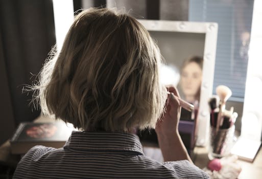 Back view of a woman applying makeup in front of a vanity mirror with lights.