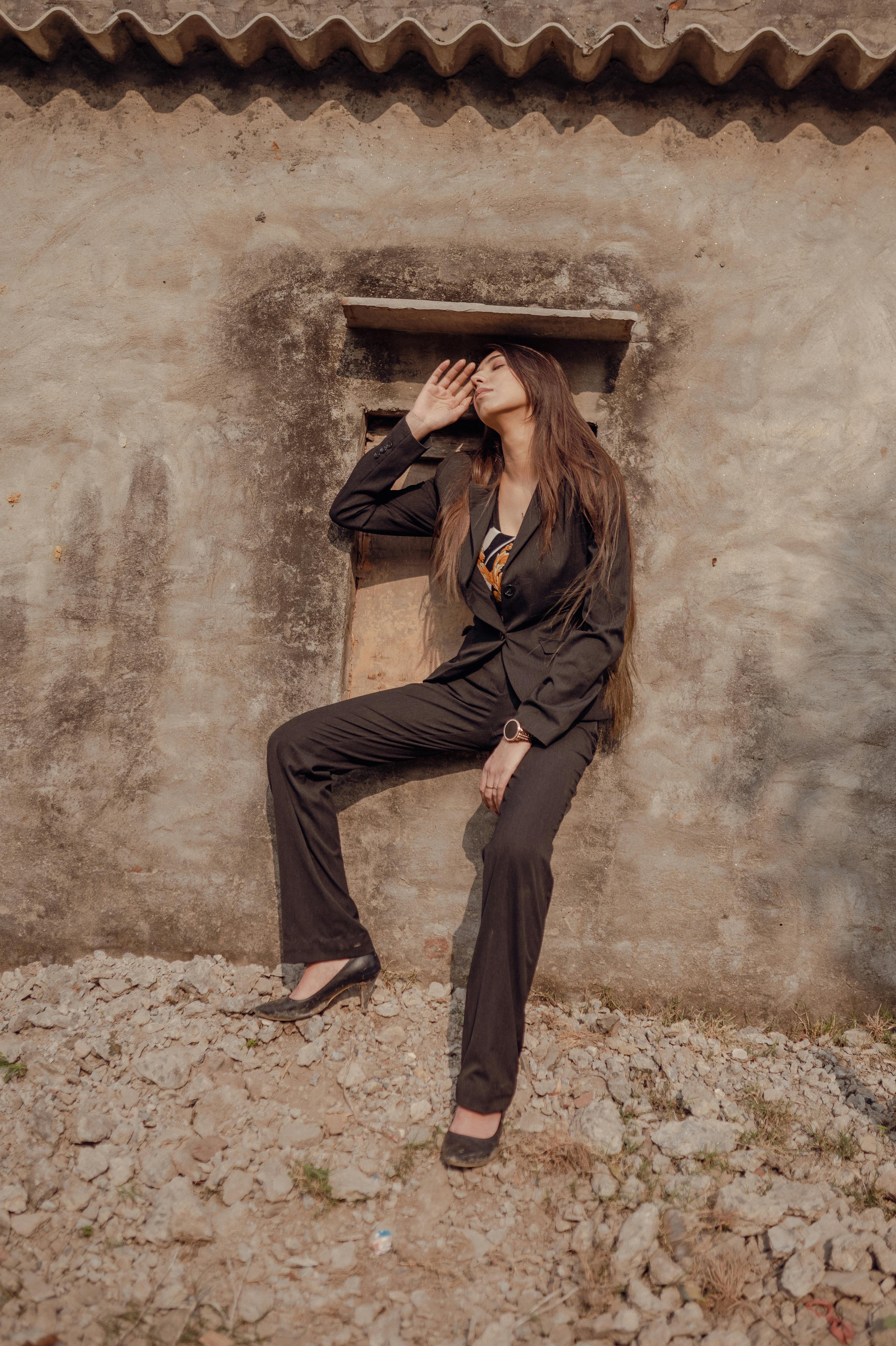 Fashionable woman in a suit poses against a rustic wall, conveying elegance.