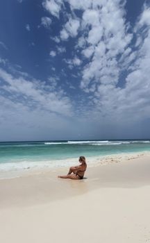 A woman in swimwear sits peacefully on a pristine beach under a clear sky, enjoying a summer retreat.
