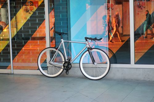 Free Silver fixed gear bike parked against a colorful window display on a city sidewalk. Stock Photo