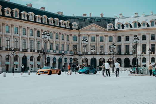 Luxury sports cars parked in a historic European plaza with grand architecture and street lamps.