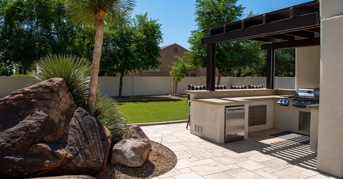 Stylish outdoor kitchen on a sunny patio with lush landscaping and clear blue sky.