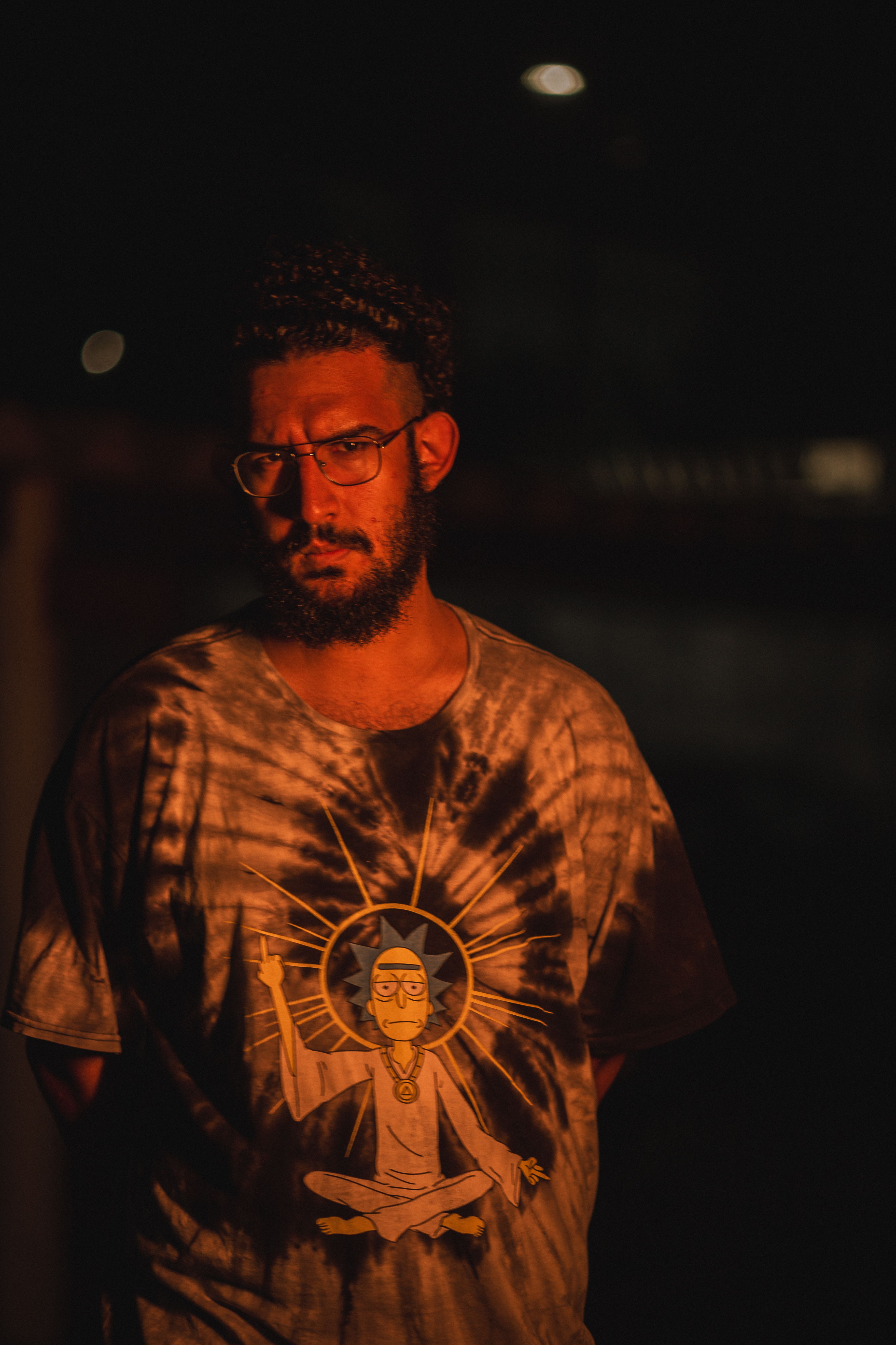 A bearded man with glasses stands in dim lighting wearing a graphic t-shirt. Captured indoors at night.