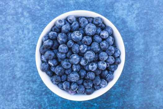 Top view of fresh blueberries in a white bowl on a textured blue background.