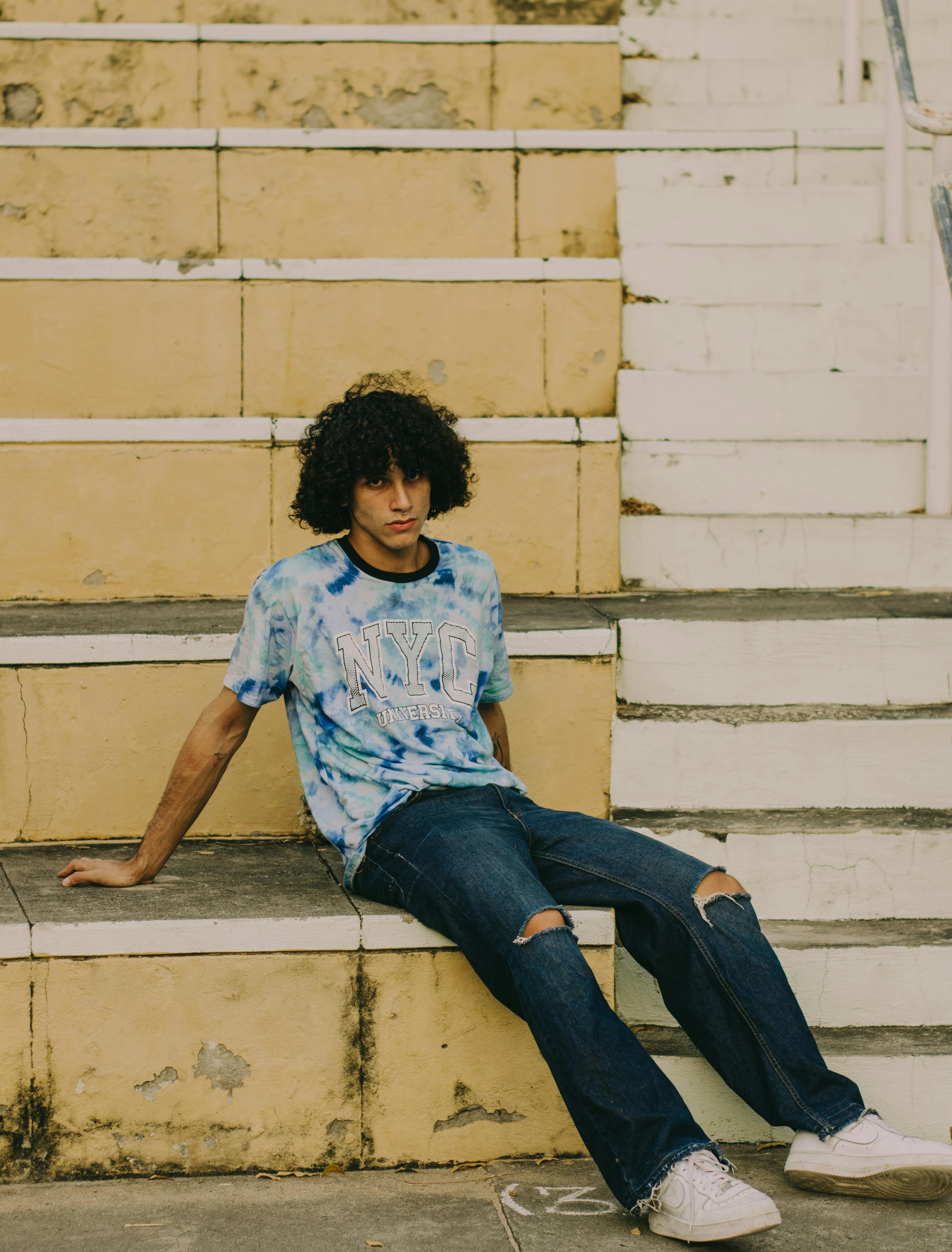 Relaxed young man with curly hair sits on urban steps in casual jeans and t-shirt.