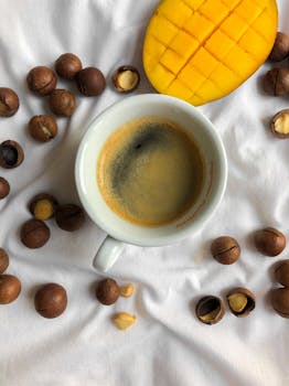 Close-up of coffee cup with sliced mango and nuts on a white surface.