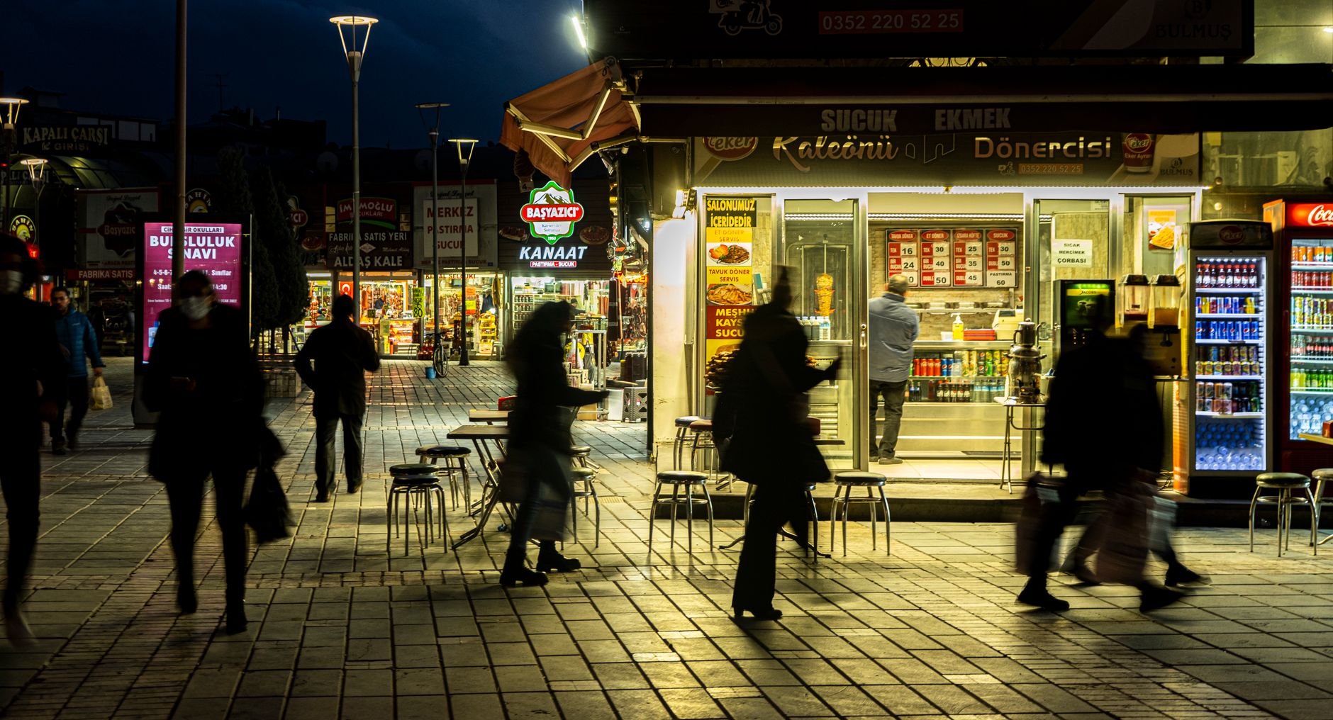 Nighttime view of a bustling street market with silhouettes of pedestrians and illuminated storefronts.