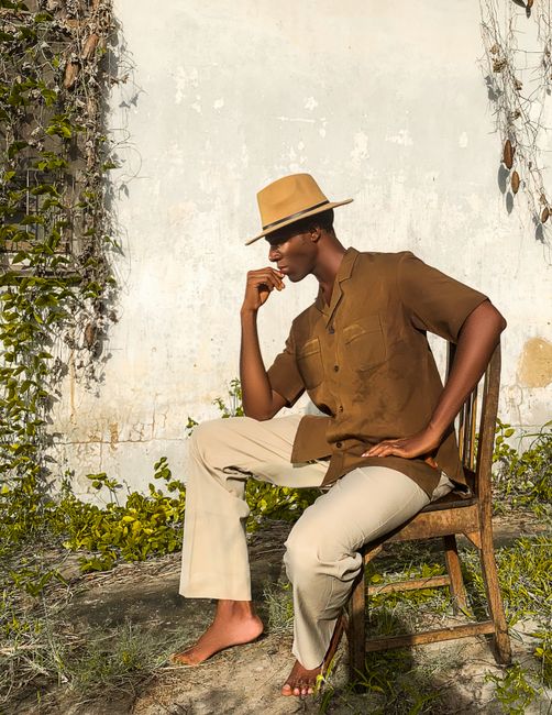A fashionable man in a hat poses elegantly outdoors against a rustic wall in Lagos, Nigeria.