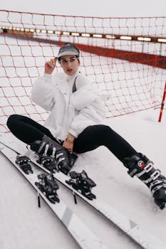 Woman in winter gear resting on snow beside skis, showcasing sports fashion and active lifestyle.