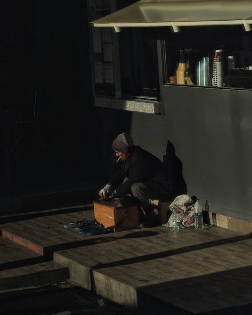 Man in Black Jacket Sitting on Sidewalk Cleaning Shoes
