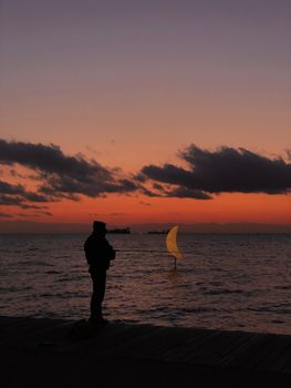 A mesmerizing silhouette of a person fishing against the sunset on Thessaloniki's seashore.
