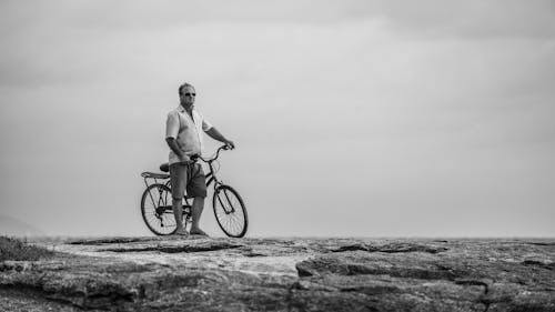 Free Grayscale Photo of Man Standing on Rocky Surface with a Bicycle Stock Photo