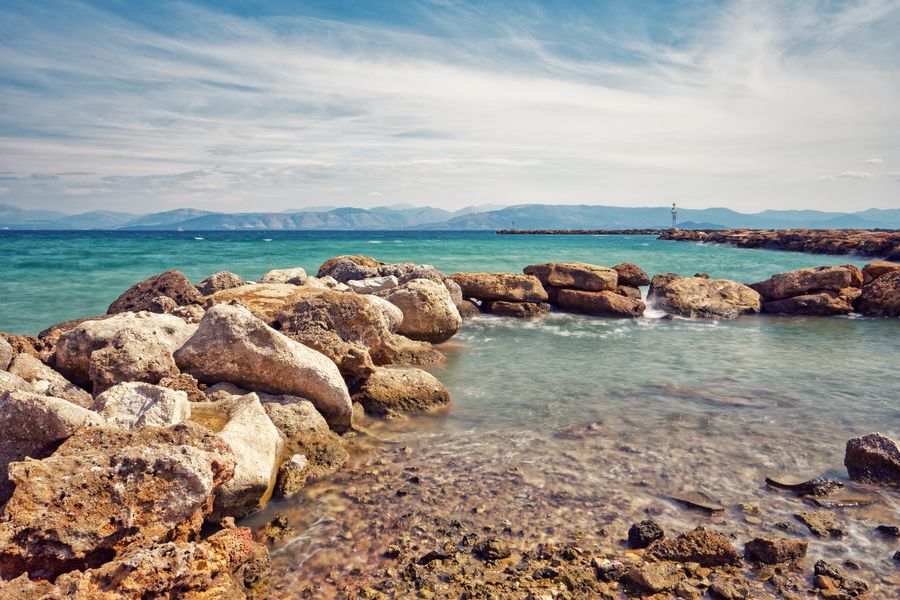 Scenic view of a rocky shoreline with turquoise waters and distant mountains under a clear sky.