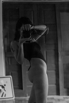 A woman in lingerie takes a black and white self-portrait with a camera indoors.