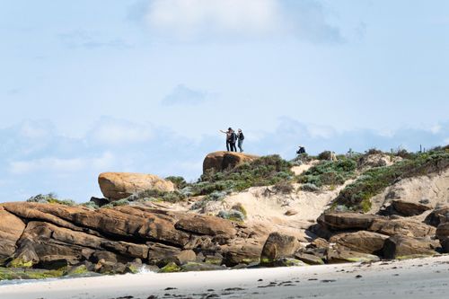 Free Two hikers standing on a rocky cliff pointing towards the ocean under a blue sky. Stock Photo