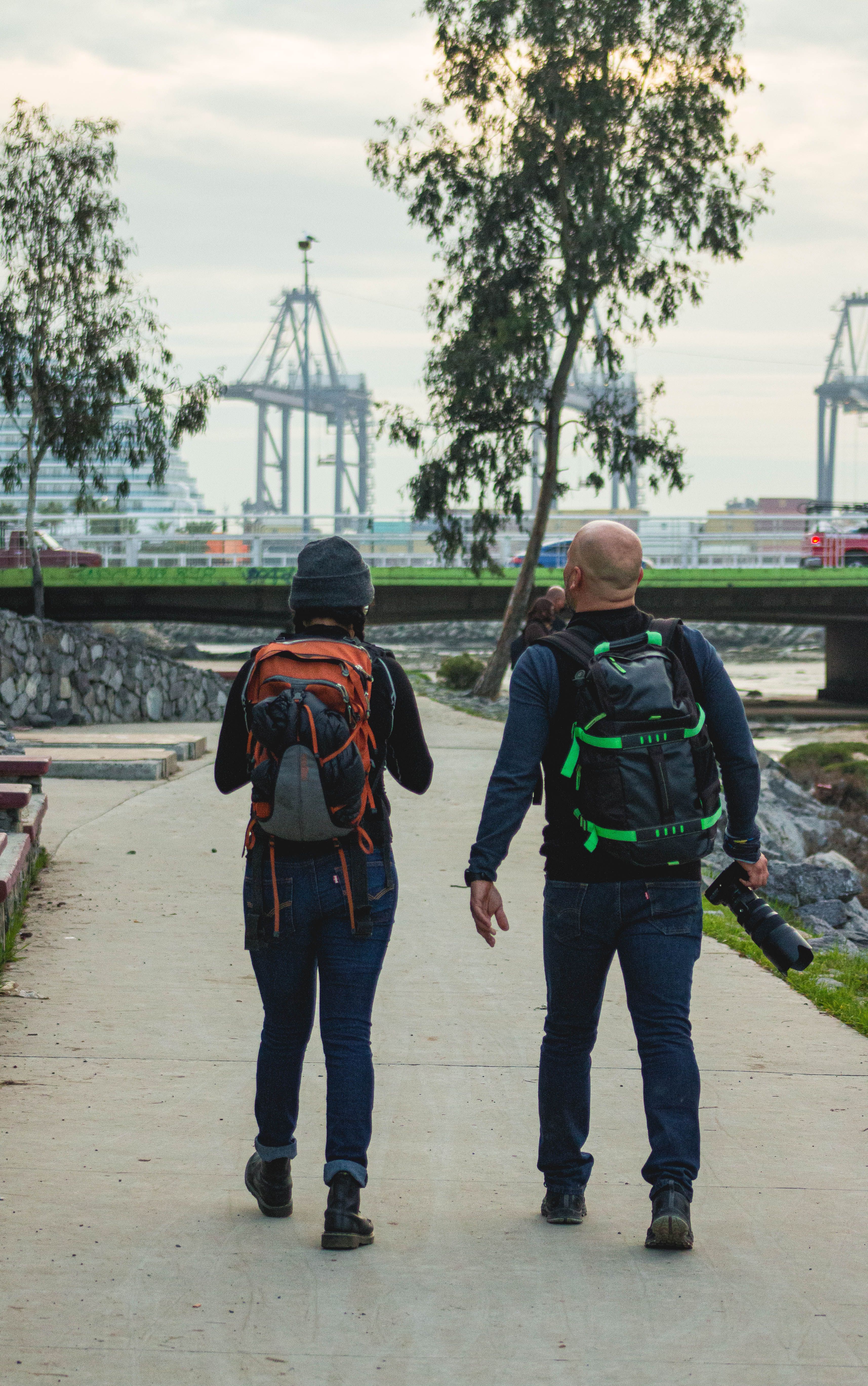 Two photographers with backpacks walking beside an urban river with industrial structures.