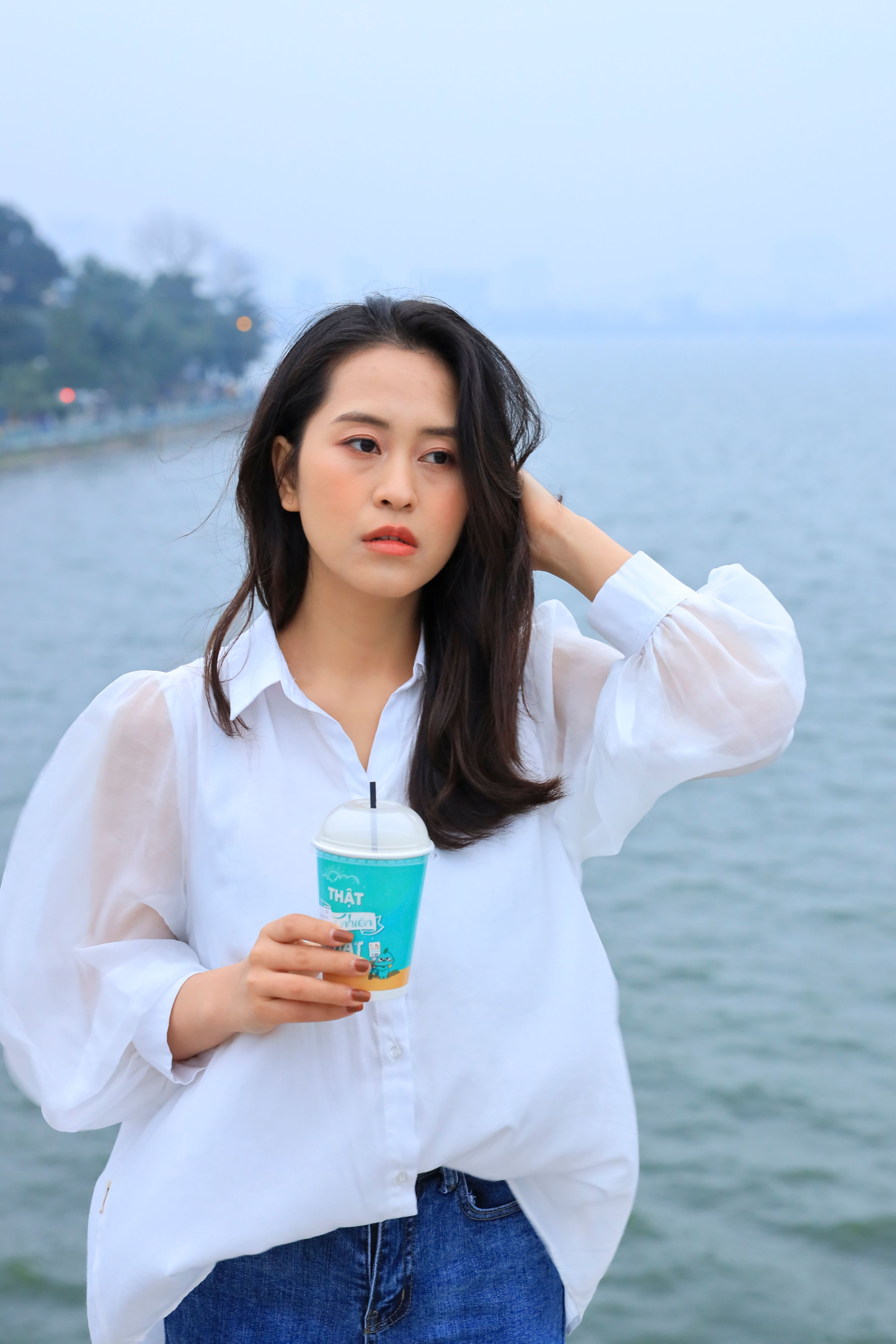 Woman in white top holding a cup by a serene waterfront, thoughtful mood.