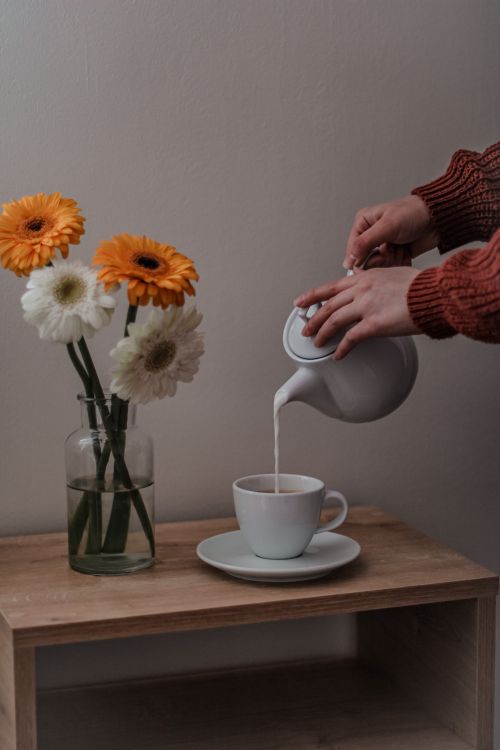 Free Hands pour milk into a coffee cup next to vibrant gerbera daisies in a vase. Stock Photo
