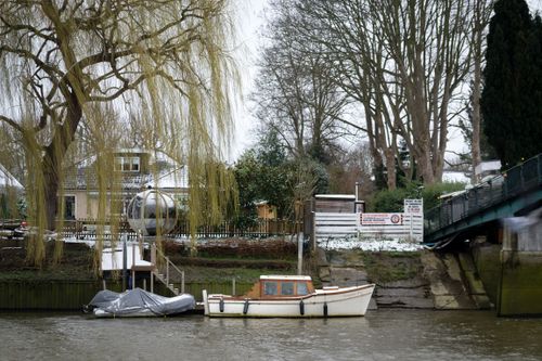 Free Scenic riverside with moored boat, willow tree, and quaint houses in Twickenham, England during winter. Stock Photo