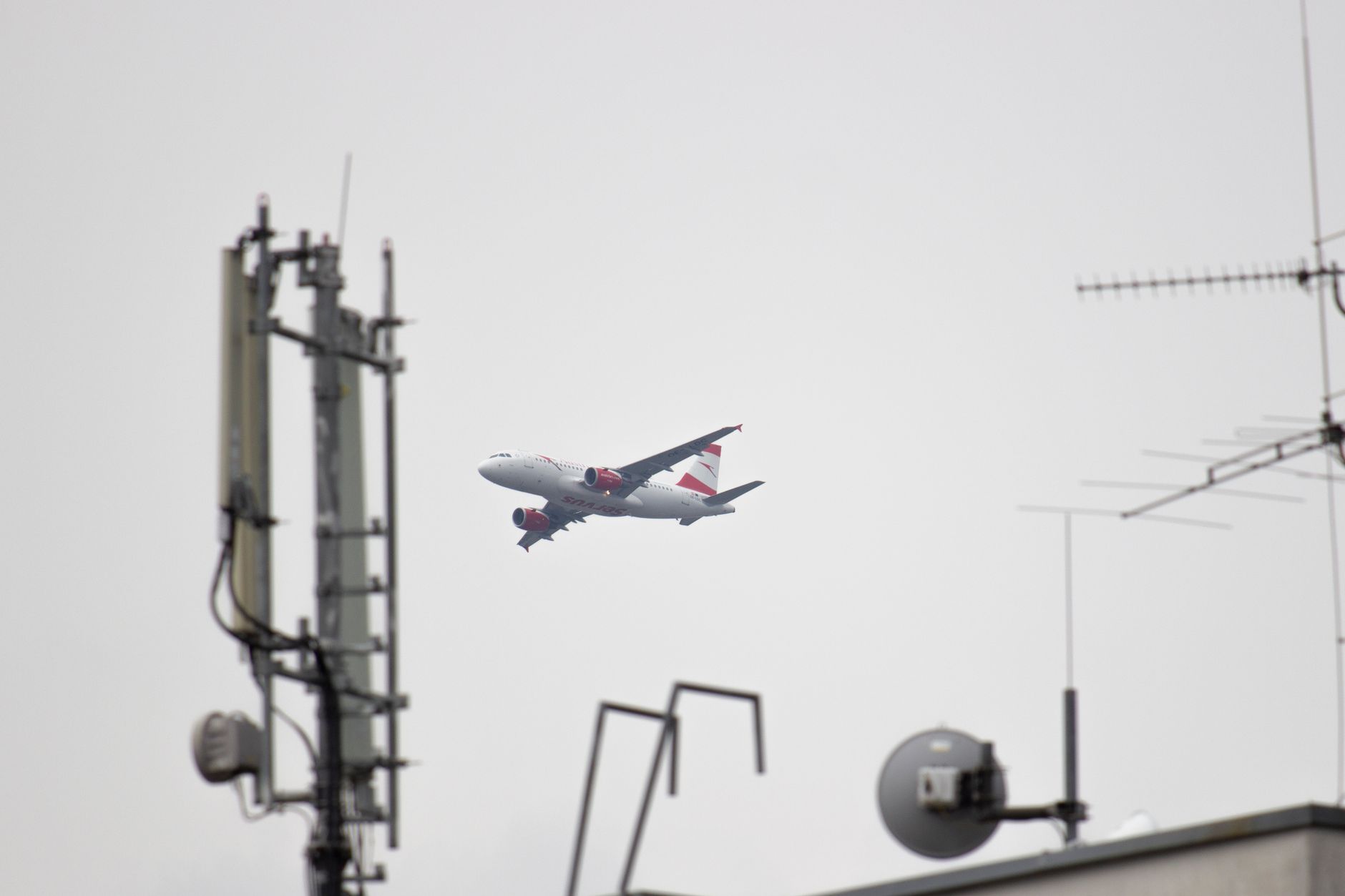 A large airliner is seen from a city rooftop with antennas, captured in daylight.