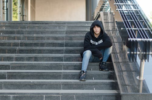 A Man in Black Hoodie and Blue Denim Jeans Sitting on Gray Concrete Stairs
