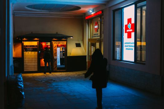 A person stands at a coffee stall beside a lit pharmacy in Saint Petersburg at night.