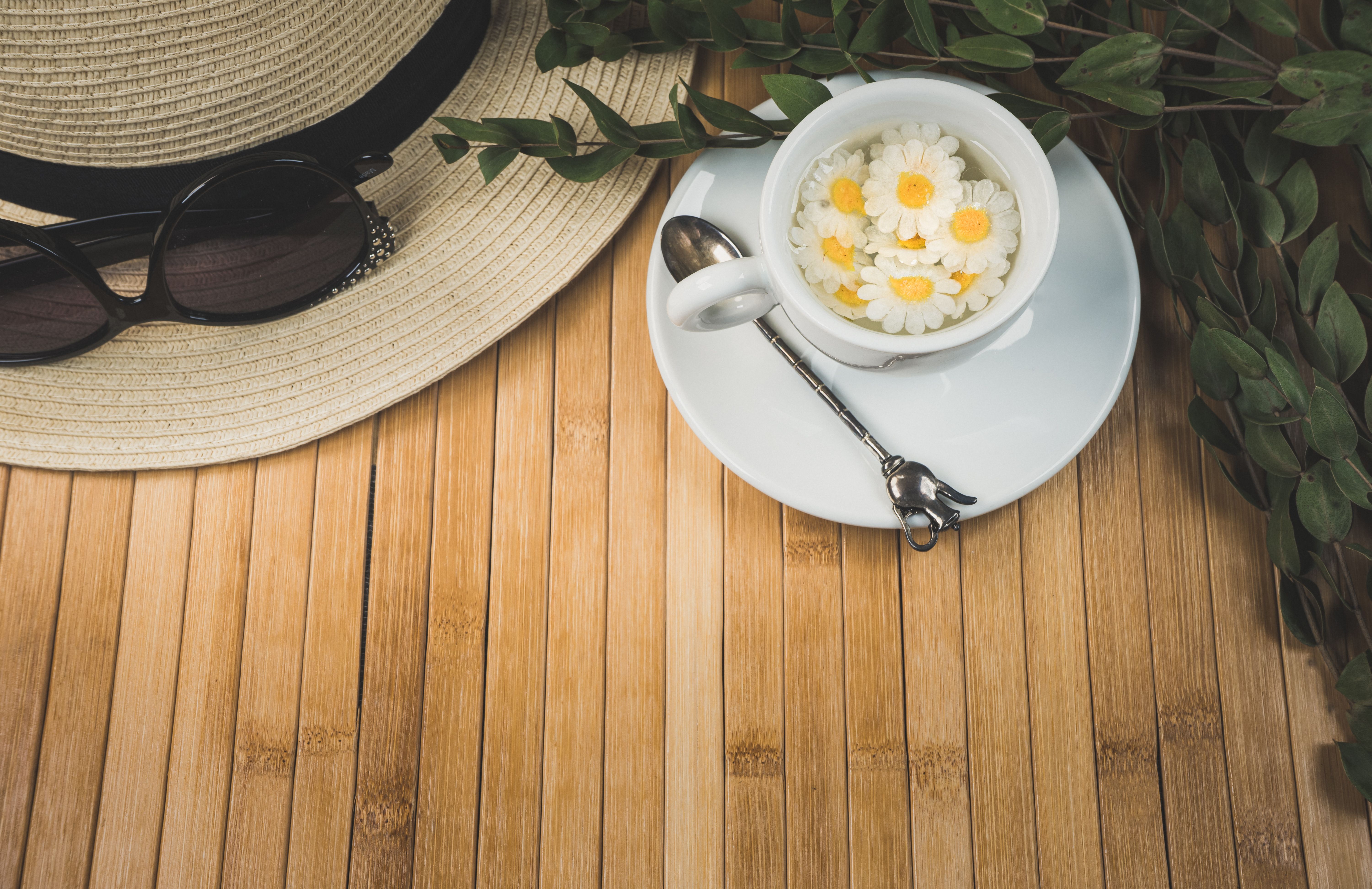 Top view of a stylish tea setup featuring a cup of chamomile flowers, hat, and sunglasses on a bamboo mat.