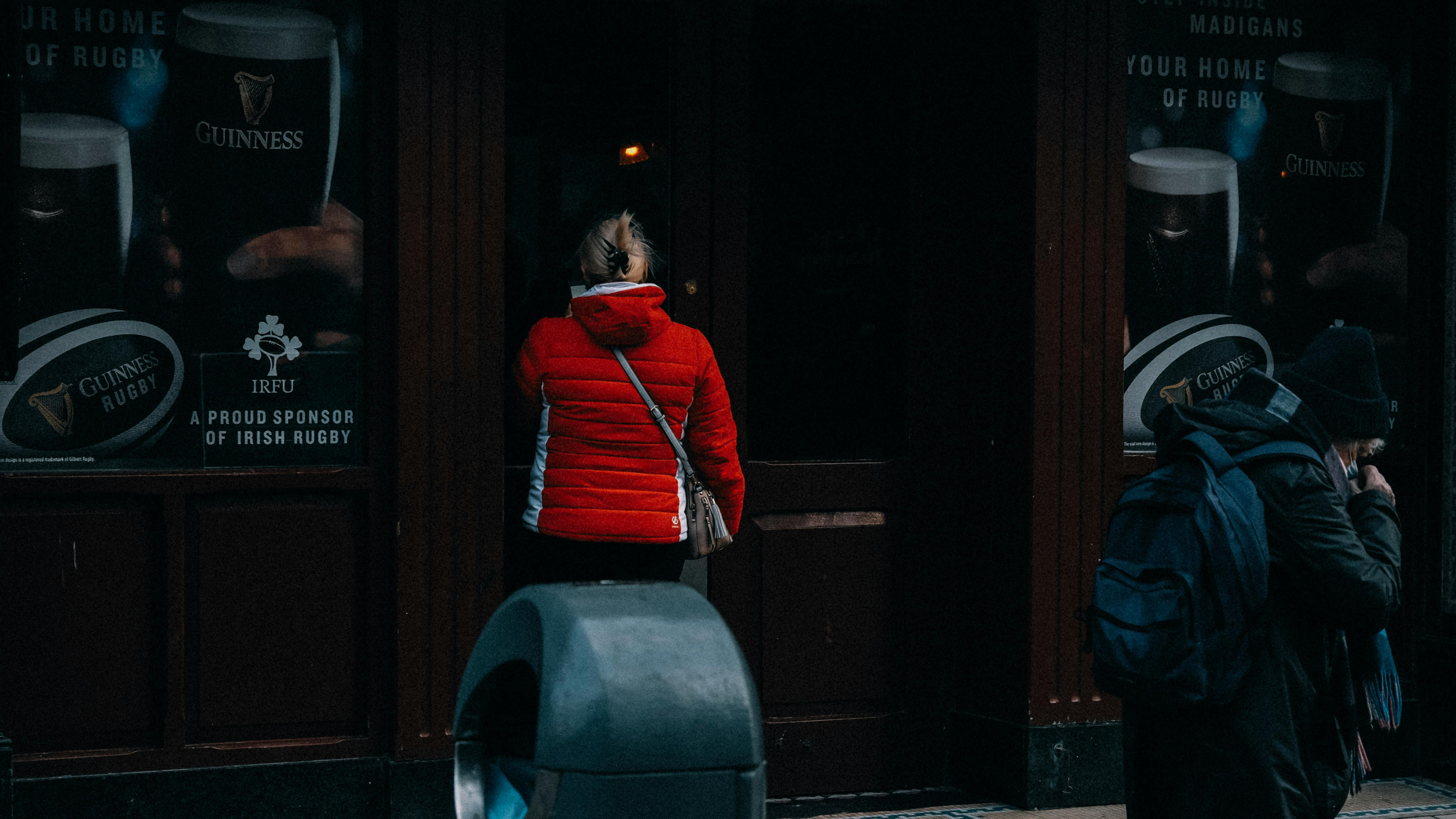 Free A woman in a red jacket stands at a pub door, viewed from behind on a city street. Stock Photo