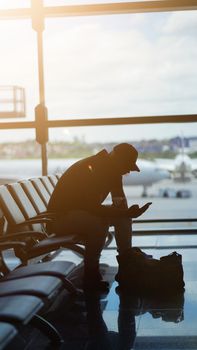 Silhouette of a man using a smartphone in an airport waiting area with airplanes visible outside.