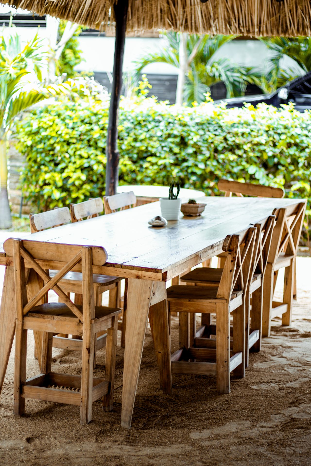 Light wood dining table with cross-back chairs under a thatched roof outdoors