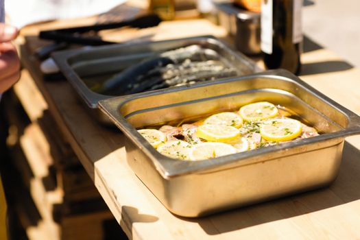 Close-up of marinated fish with lemon slices and herbs in stainless steel trays outdoors.
