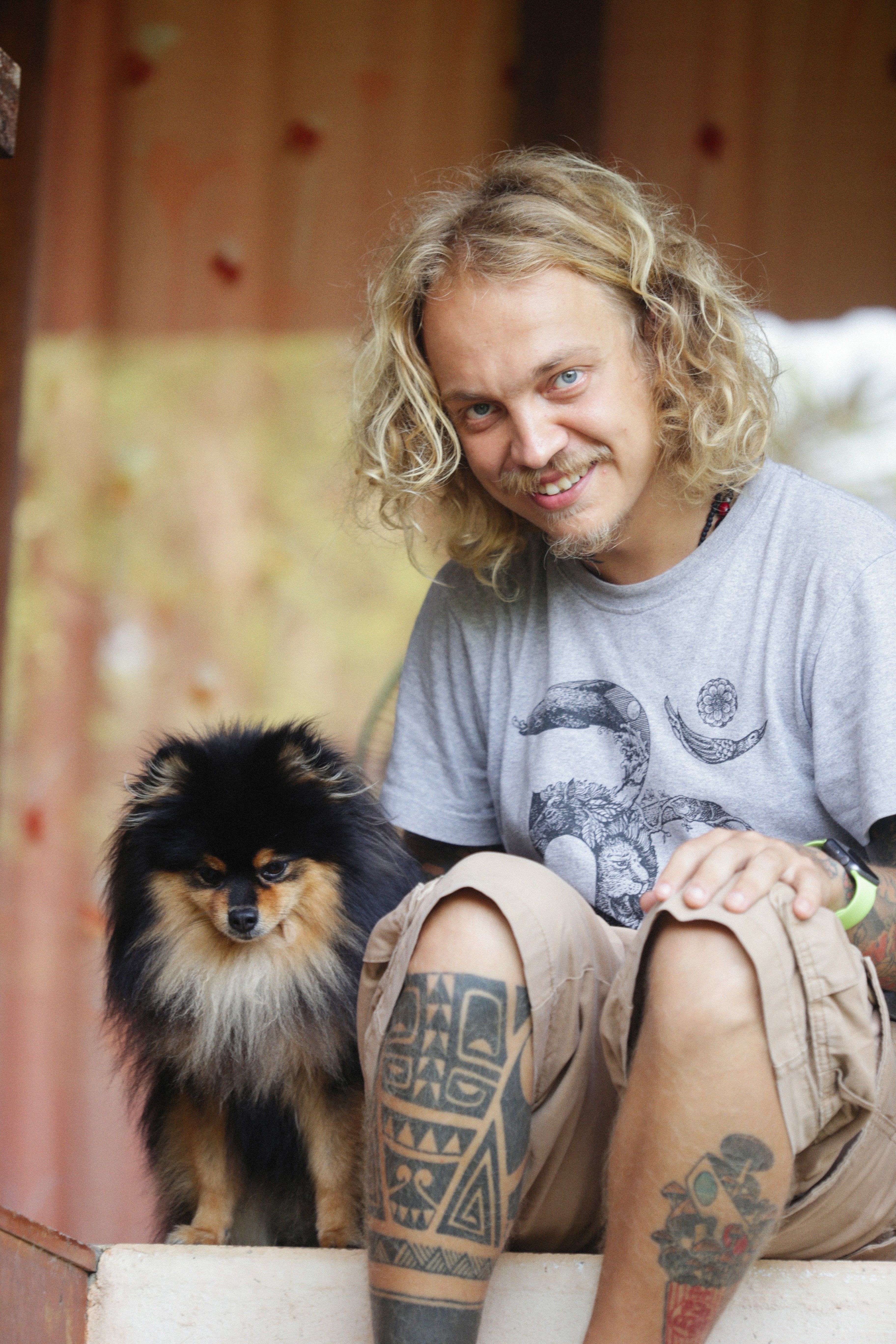A man with tattoos smiling alongside a cute Pomeranian dog indoors.