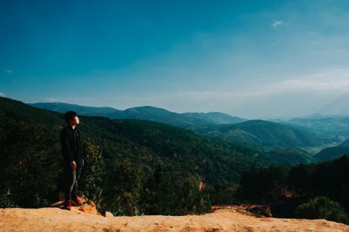 Free A man stands on a hilltop overlooking a vast, scenic mountainous landscape under clear daylight skies. Stock Photo