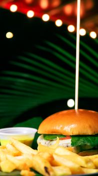 Delicious cheeseburger served with crispy fries under palm leaves at night.