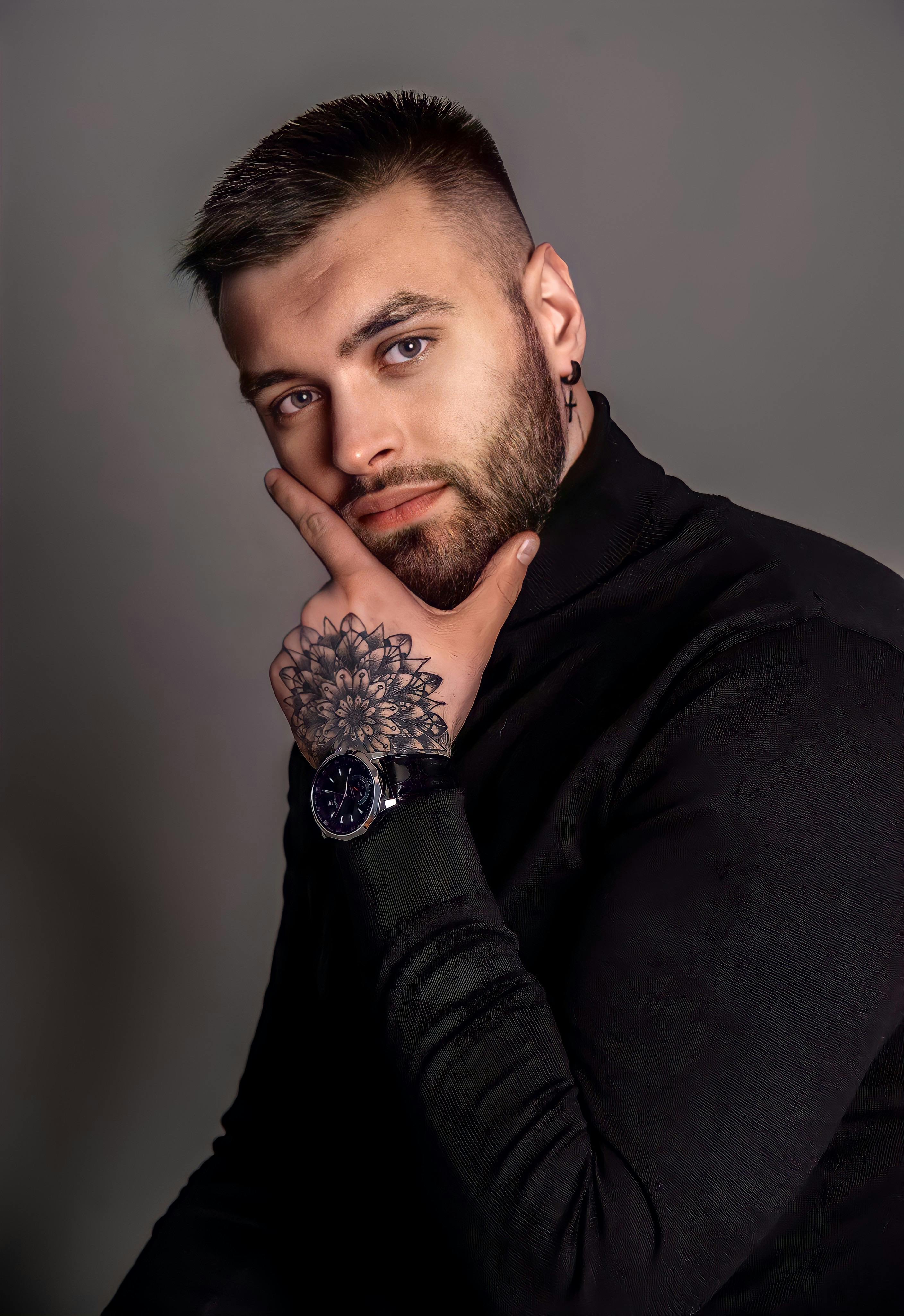 Close-up portrait of a young man with a beard and tattoo in stylish pose.