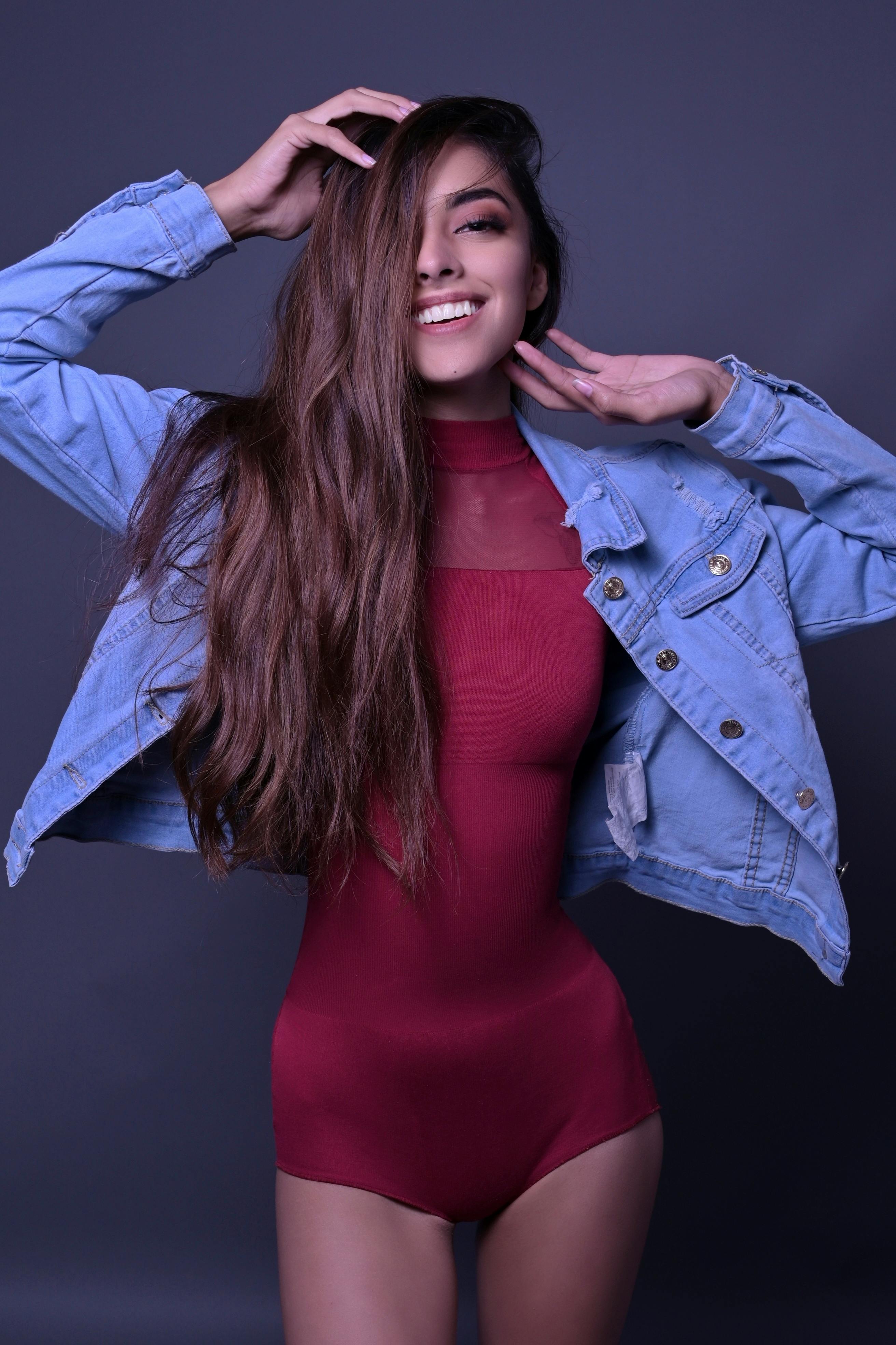 Confident woman in denim jacket and red bodysuit posing against a dark background.