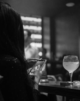 A woman using a smartphone in a dimly lit bar with a cocktail on the table.