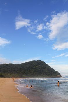 A serene beach scene with people swimming and a lush hill in the background under a clear sky.