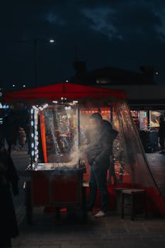 Urban street food vendor grilling at night under a red stall canopy, lights create a cozy atmosphere.