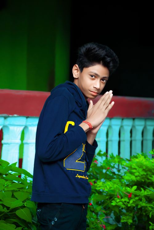Free A teenage boy in a blue hoodie makes a greeting gesture outdoors, surrounded by greenery. Stock Photo