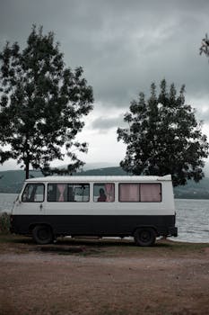 A vintage van sits beside a tranquil lake, surrounded by trees, under a cloudy sky.